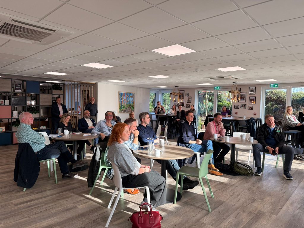 A side on view of a Group of people sitting at tables and chairs looking towards the front of a room, listening